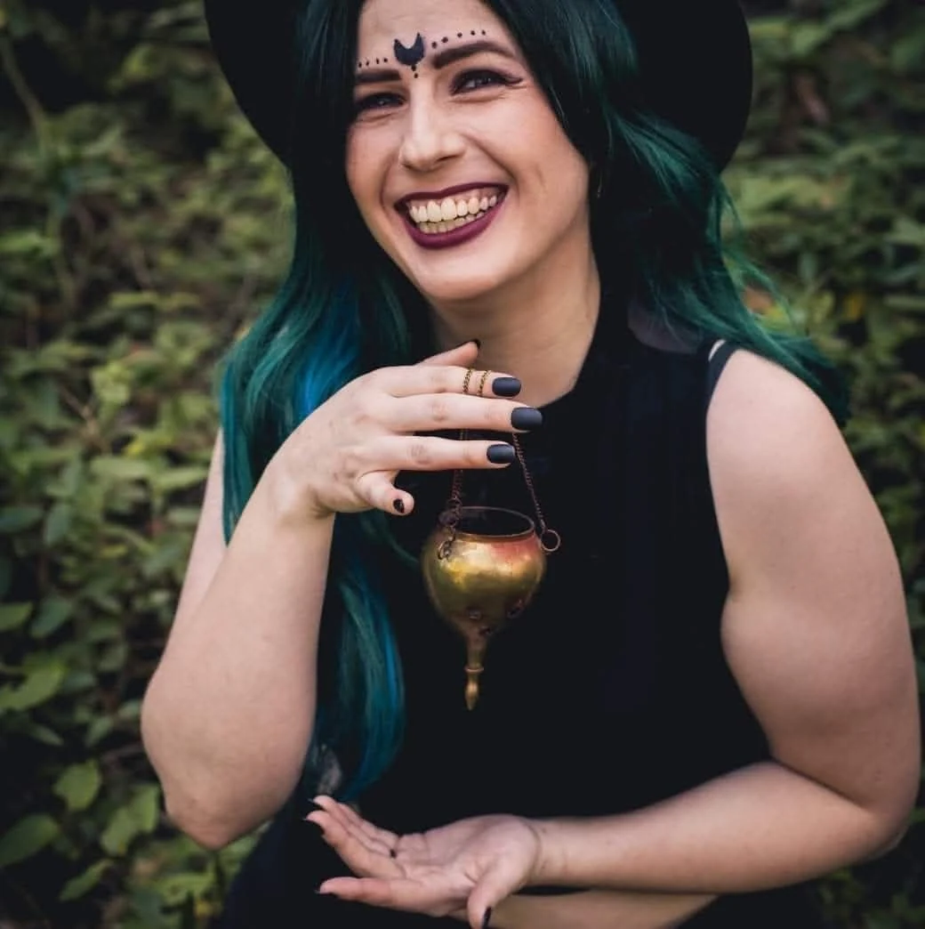 Alannah smiling in a black hat, with long teal hair, in a wooded setting